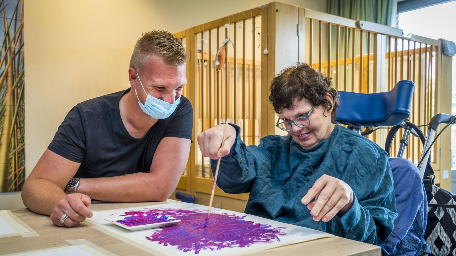 Gehandicaptenzorg: begeleider helpt cliënte met schilderen op papier